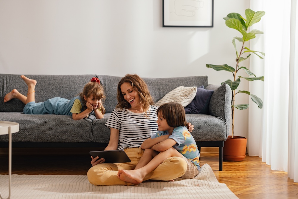 A woman sitting on the floor with two children in a living room, looking at a tablet and smiling.