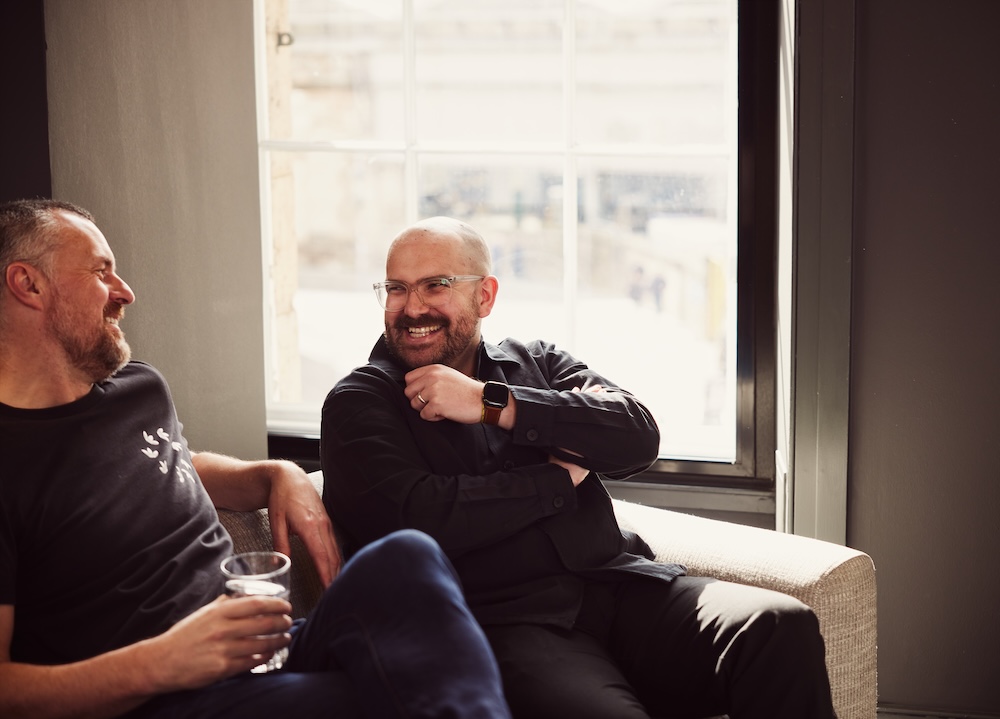 Two men sitting on a sofa in a well-lit room, engaging in a casual conversation and sharing a laugh. The overall mood of the image is friendly and relaxed.