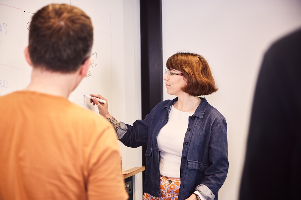 A man and a woman standing in front of a whiteboard. The woman is writing notes on the board with a marker.